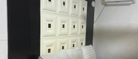 A hotel room with a white bed, a black nightstand with a phone, a white headboard, and an air conditioning unit on the wall.