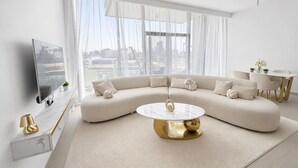 A modern living room with a large curved sofa, a round coffee table, a TV on a stand, and a dining area with a table and chairs. - 1 bedroom in new building by luxury developer (Dubai)