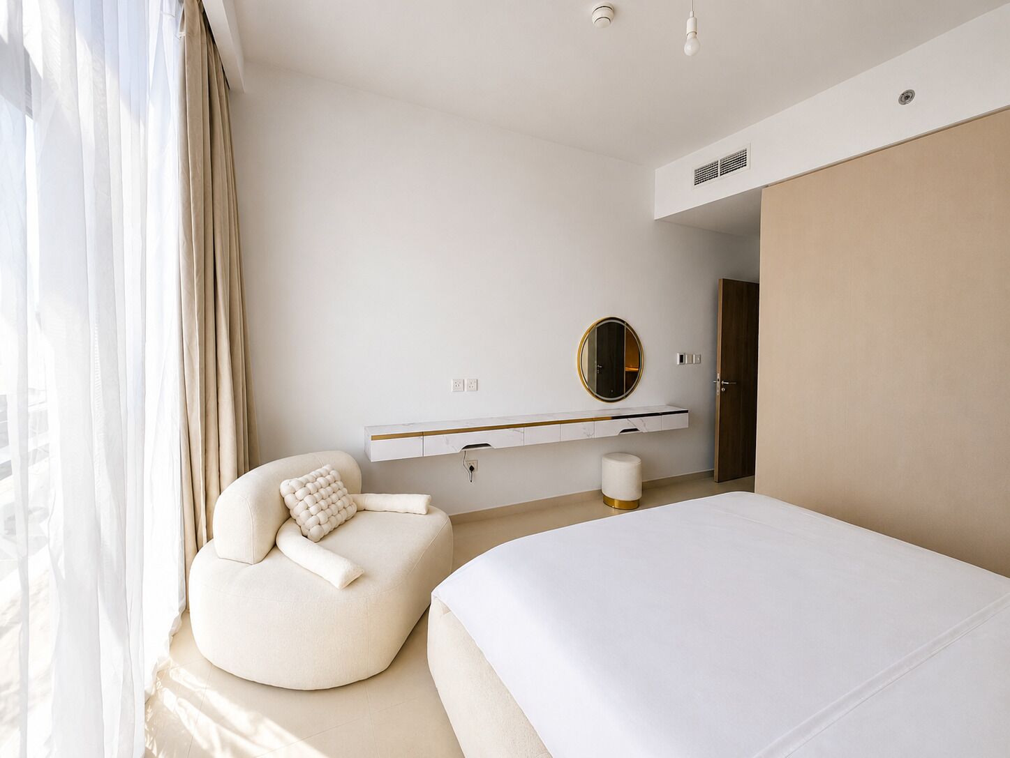 A modern hotel room with a large bed, a round mirror, a sofa with a textured pillow, and a small side table.