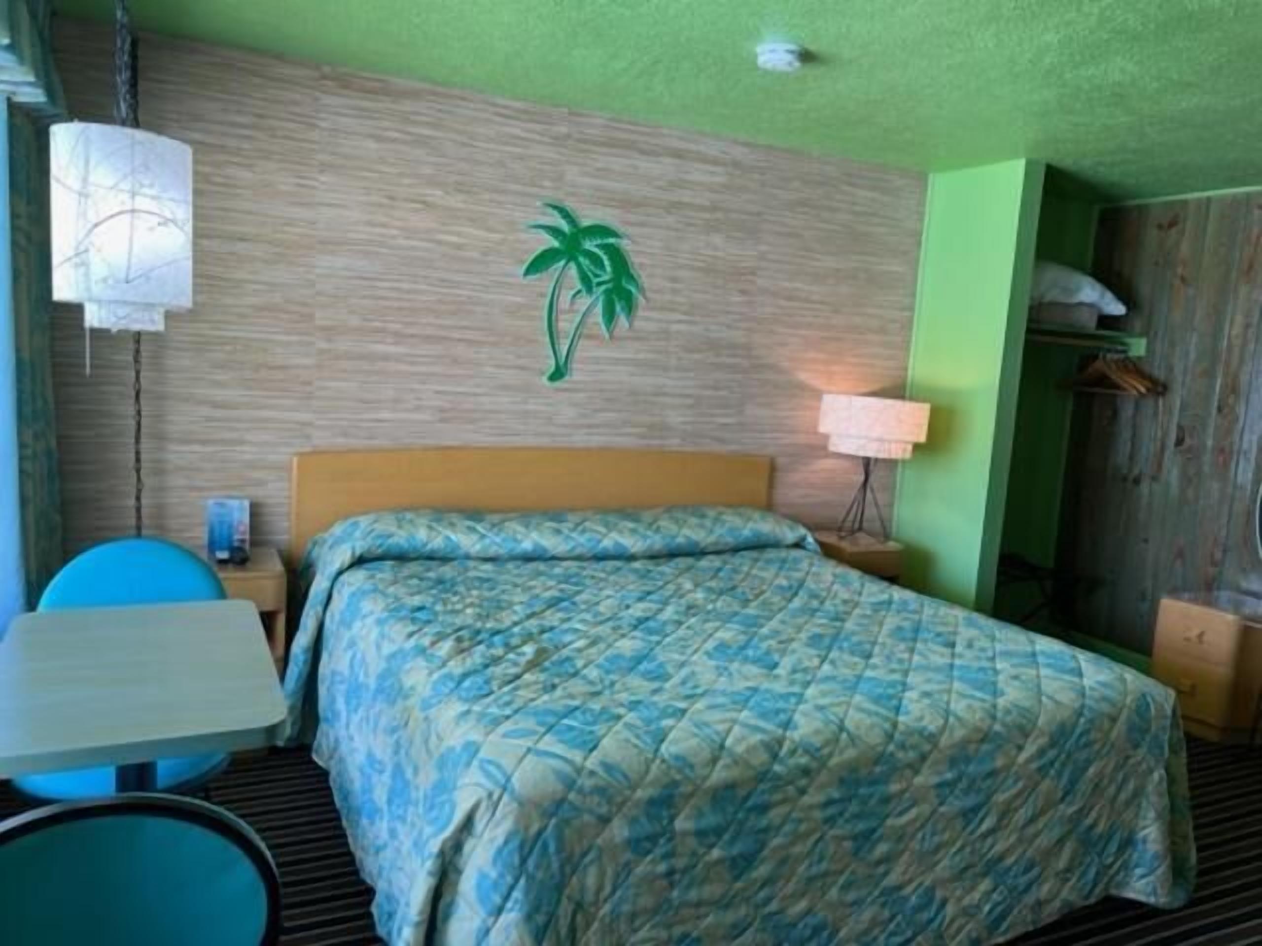 Habitación de hotel con cama, una mesa, una silla, una lámpara y una planta en la pared.