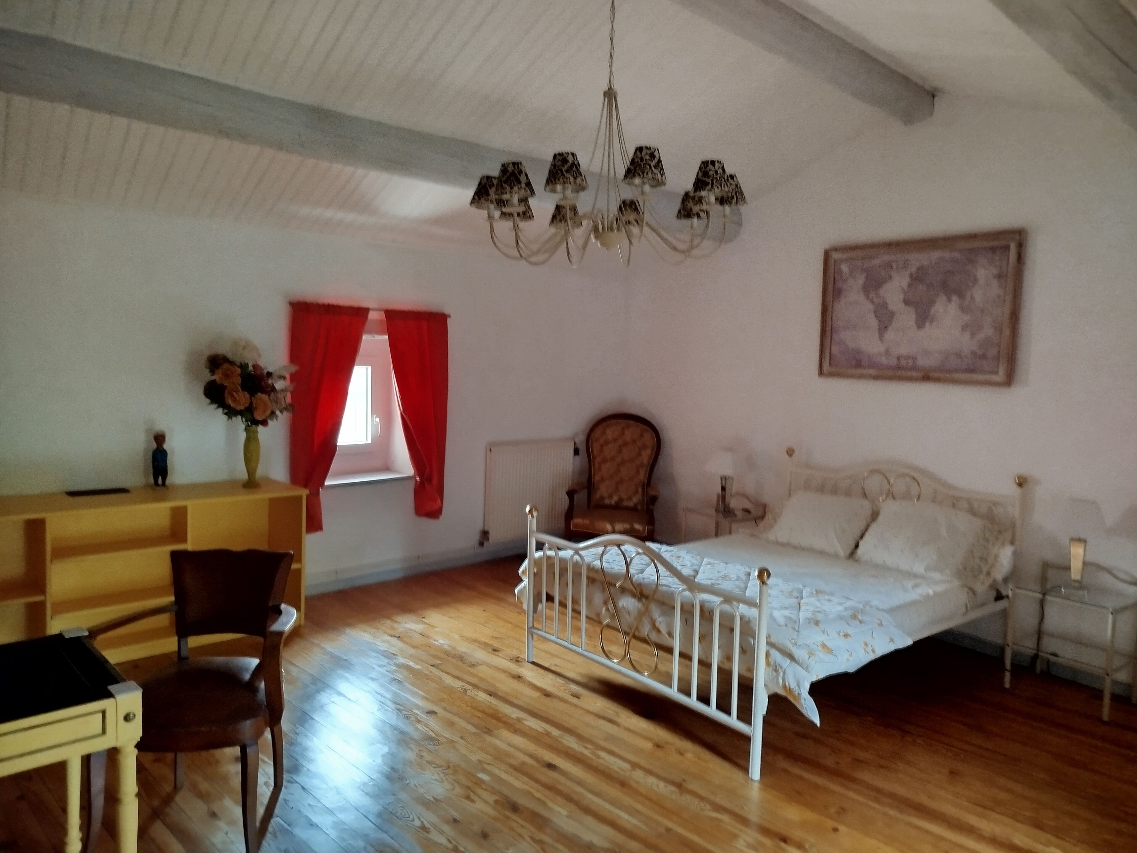 Un dormitorio con suelo de madera, una cama blanca, una silla, un escritorio, una cortina roja, una araña y un mapa del mundo en la pared.