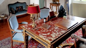 Une pièce avec un lustre, un escalier, un canapé bleu, une table rouge et un miroir.