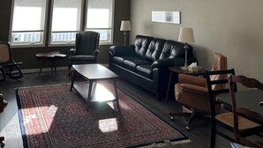 Ein Wohnzimmer mit hoher Decke, zwei Fenstern, einem Deckenventilator, einer schwarzen Ledersofa, einem Holztisch und einem gemusterten Teppich.