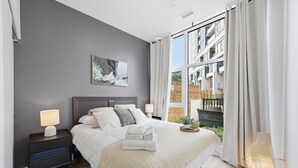 A modern bedroom with a large bed, a nightstand, a bedside lamp, a window with curtains, and a city view.