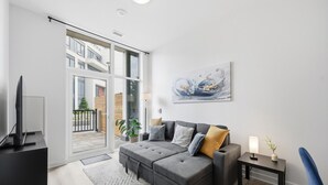 A modern living room with a grey sofa, a flat-screen TV, a painting, and a view of a building through large windows.
