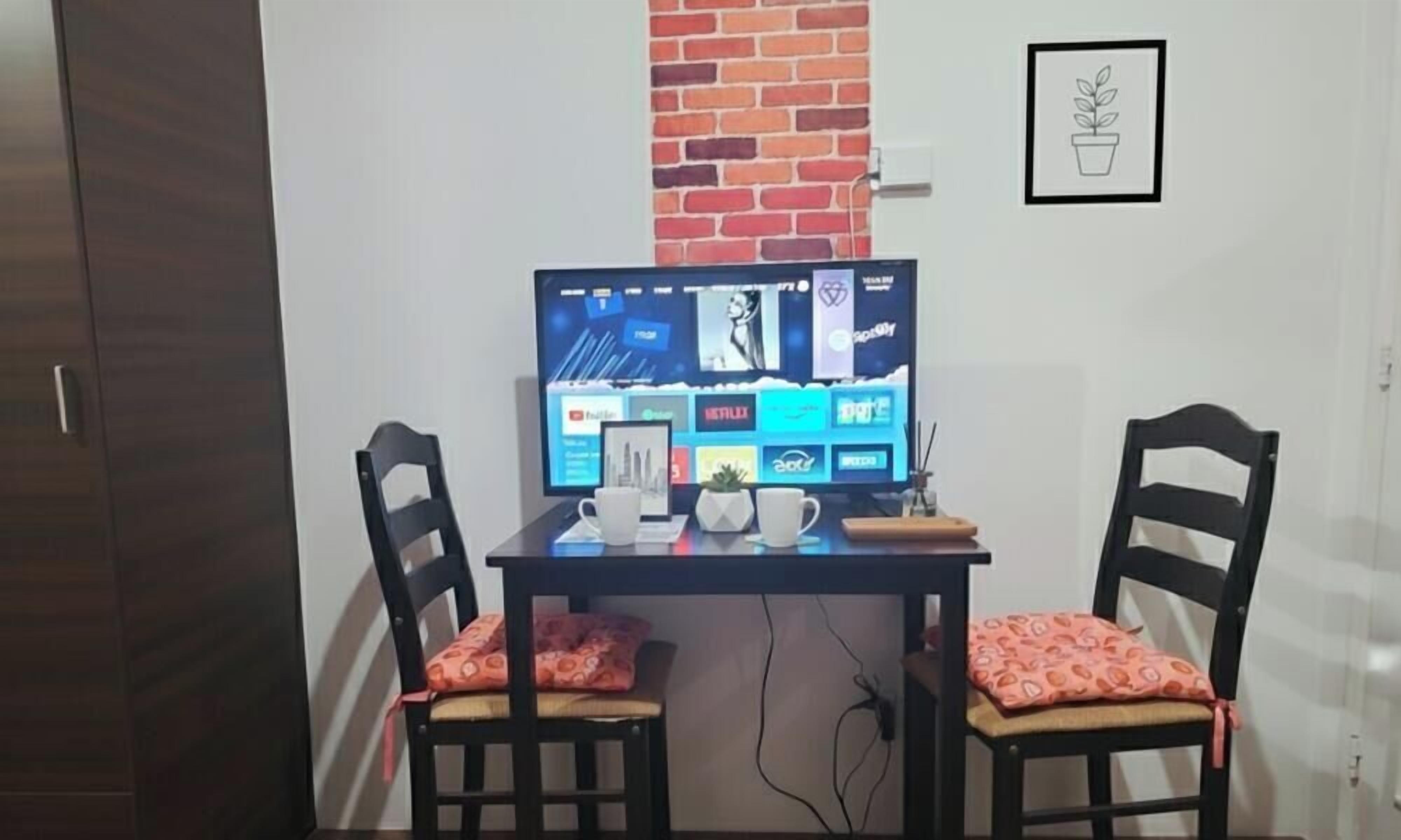 Un espacio de trabajo con un monitor de computadora mostrando servicios de transmisión, dos sillas con cojines, una mesita con una taza y una planta, y una pared de ladrillo.