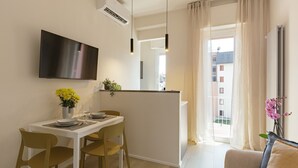 Een moderne woonkamer met een flatscreen-tv aan de muur, een eethoek met een tafel en stoelen, en uitzicht op het buitenlands gebied door een groot raam.