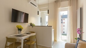 A modern living room with a flat-screen TV mounted on the wall, a dining area with a table and chairs, and a view of the outside through a large window. - Tabata Home (Milano)