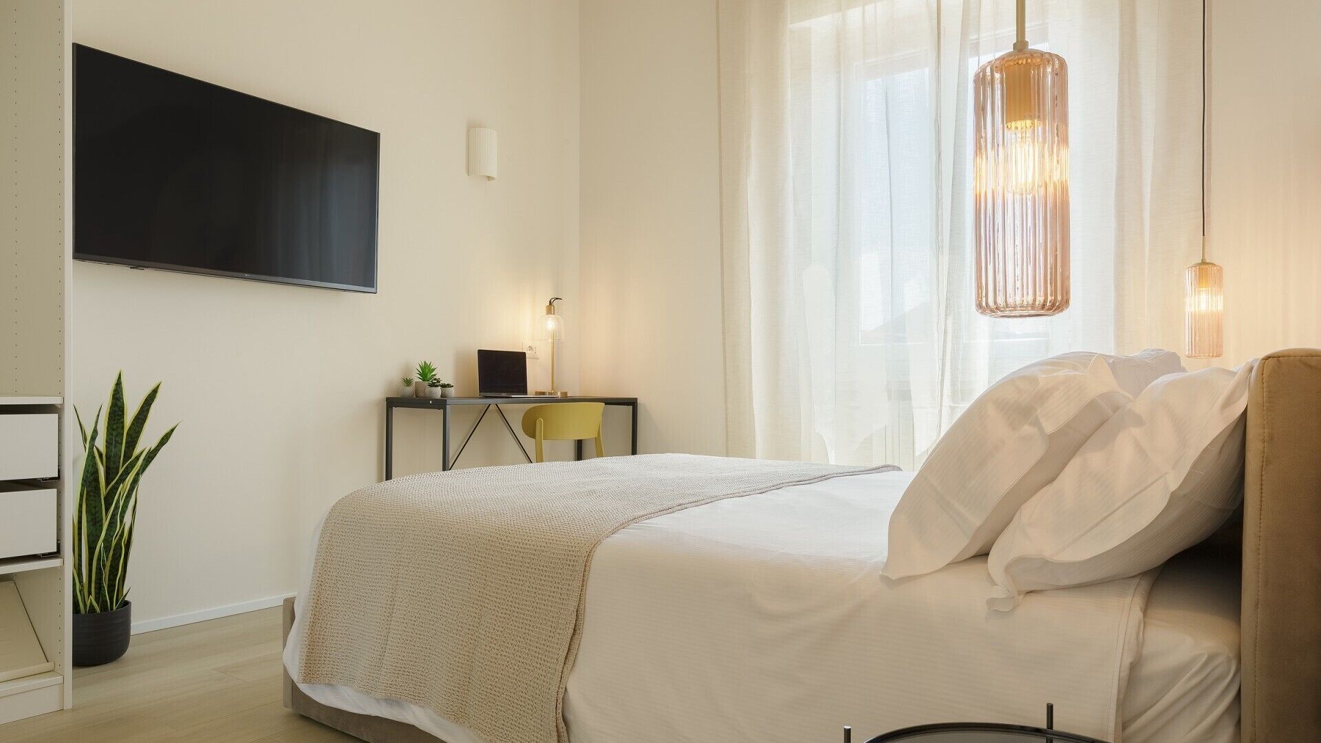A bedroom with a bed, a desk with a laptop, a TV mounted on the wall, and a hanging light fixture.