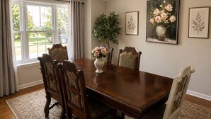 En spisestue med et stort træbord, fire polstrede stole, en vase med blomster, en lysekrone og indrammede botaniske illustrationer på væggene.