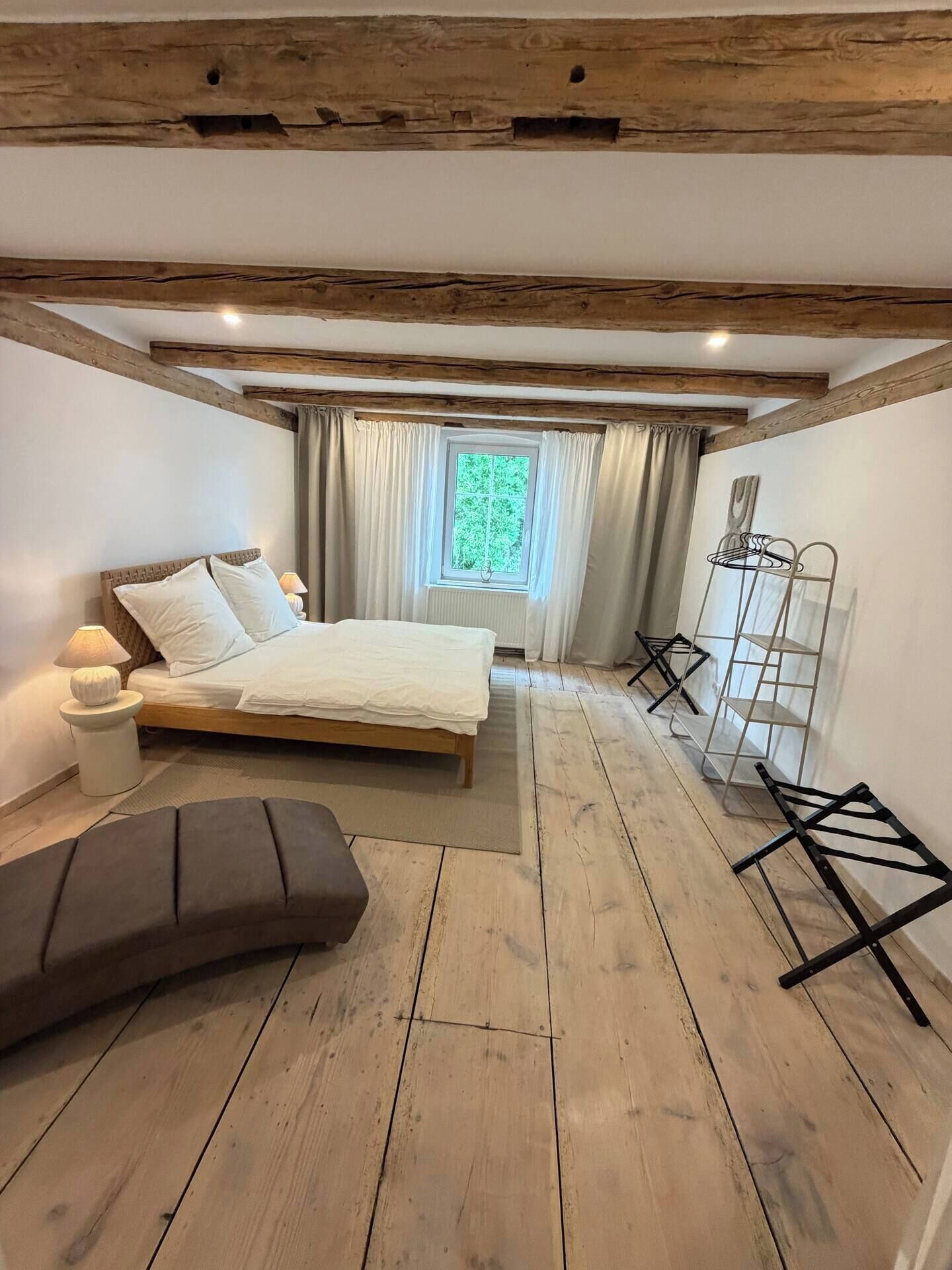 Ein Schlafzimmer mit Holzboden, einem Bett, einer Bank, einem Fenster mit Vorhängen und einem Metallregal.