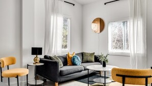 Un salon moderne comprenant un canapé, deux fauteuils, une table basse, une lampe et deux fenêtres ornées de rideaux légers.