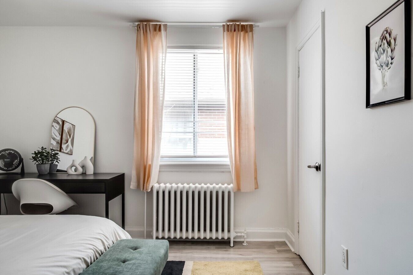 A bedroom with a bed, a desk, a chair, a radiator, and a window with curtains.