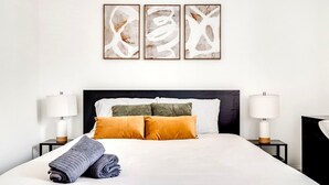 Un lit bien fait, recouvert d’une couette blanche, avec deux oreillers et une serviette enroulée. On y trouve deux tables de chevet avec des lampes, et une œuvre d’art abstraite accrochée au mur.