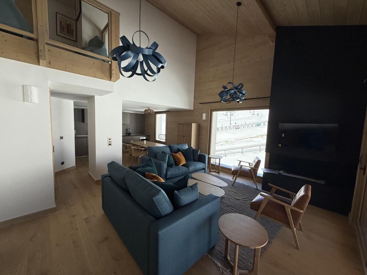 Ein modernes Wohnzimmer mit einer Couch, Couchtisch und Sesseln, ein Essbereich mit Tisch und Stühlen, eine Küche im Hintergrund und ein großes Fernsehgerät an der Wand.
