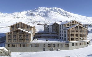 Un chalet à étages, doté d’une architecture symétrique, de grandes fenêtres et de balcons en bois, le tout se dressant sur fond de montagnes enneigées.