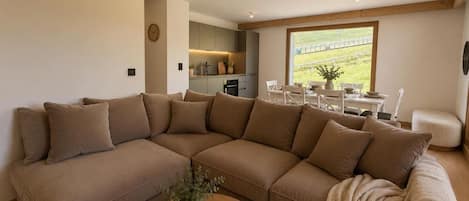 Un salón con un amplio sofá seccional en forma de L, una mesa de centro con un libro y una planta, y vistas a una cocina y zona exterior a través de un ventanal.