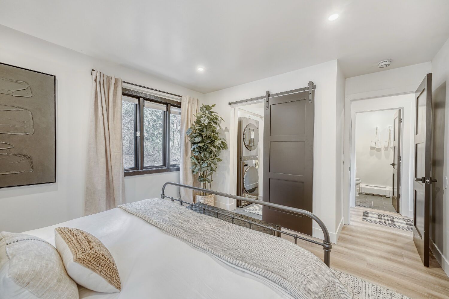 Een slaapkamer met een bed, een raam, een plant, een spiegel en een deur naar een badkamer.