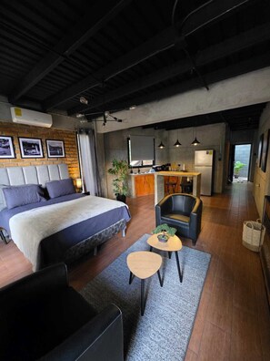Habitación de hotel moderna con una cama grande, un área de descanso con una mesita y una kitchenette al fondo.