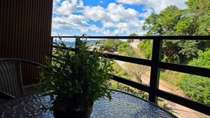 A balcony with a potted plant, a wicker chair, and a view of the sky and greenery.