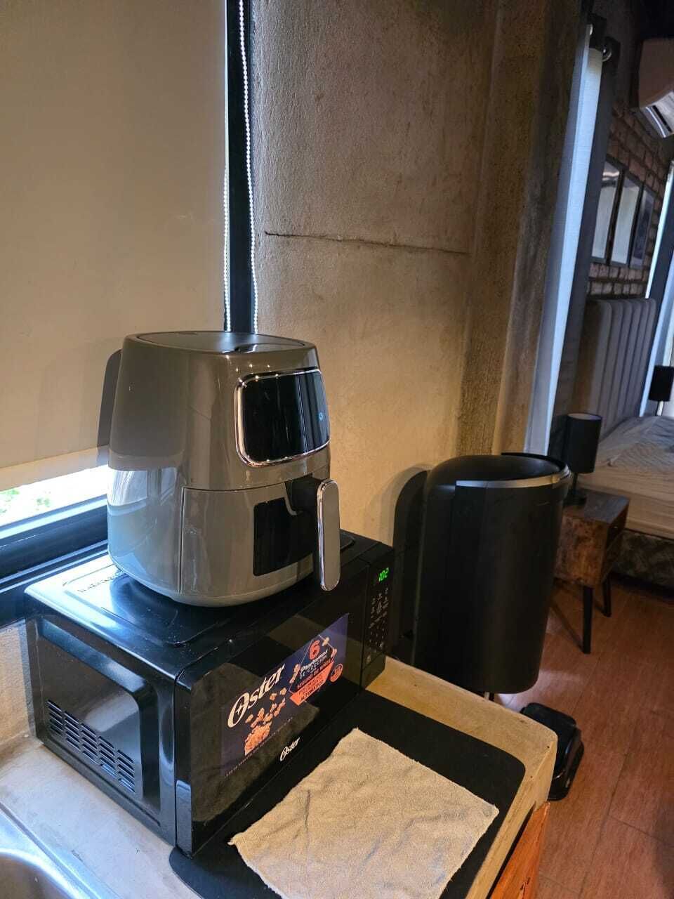 Una cafetera sobre una encimera con una toalla y un recipiente negro, probablemente para posos de café, en una habitación con una ventana y una puerta al fondo.