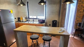 Una cocina con una isla central, dos taburetes de bar, iluminación colgante, un refrigerador y una pared de ladrillo.