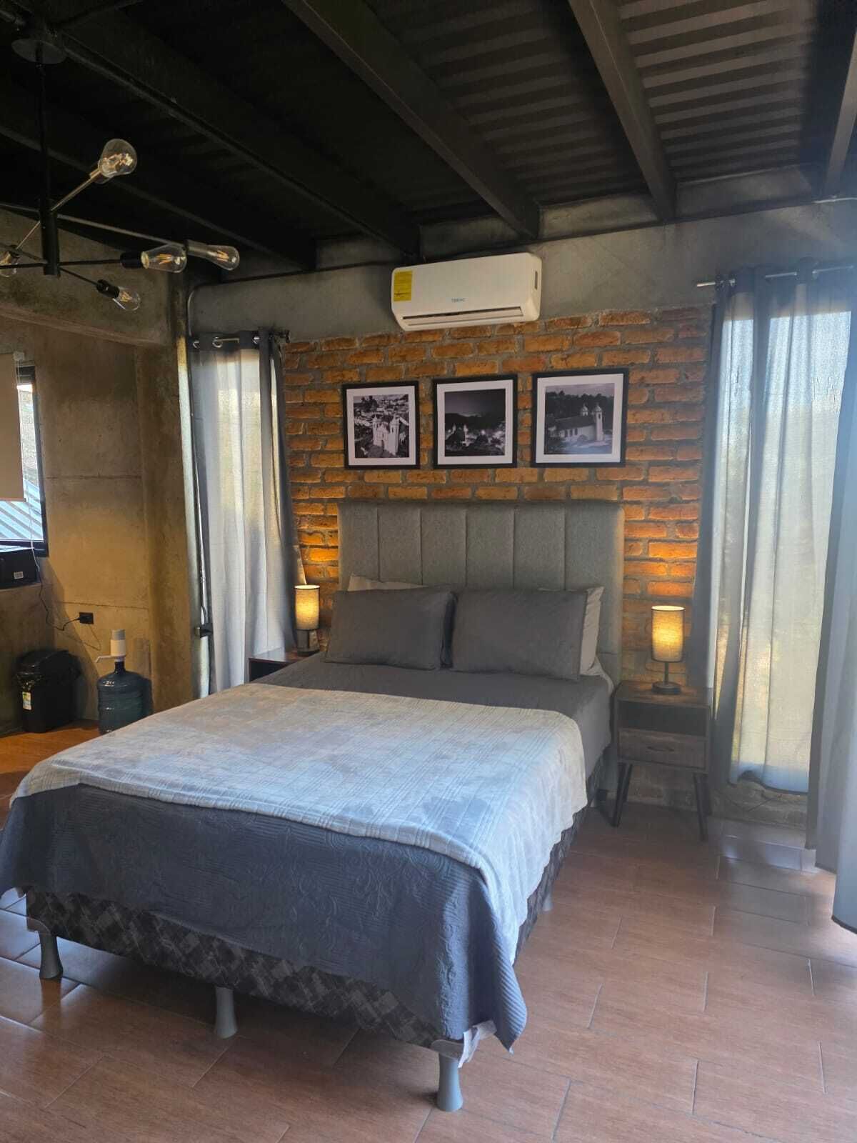 Un dormitorio con cama, cabecera, aire acondicionado de pared y ventana con cortinas.