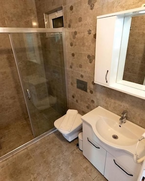 A bathroom with a glass shower enclosure, a toilet, a sink with a mirror, and tiled walls. - Doğa pansiyon (muğla)