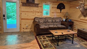 Het interieur van een blokhut met een houten wand, een opgehangen hertenhof, een bank, een bijzettafeltje en een deur met uitzicht op groen.