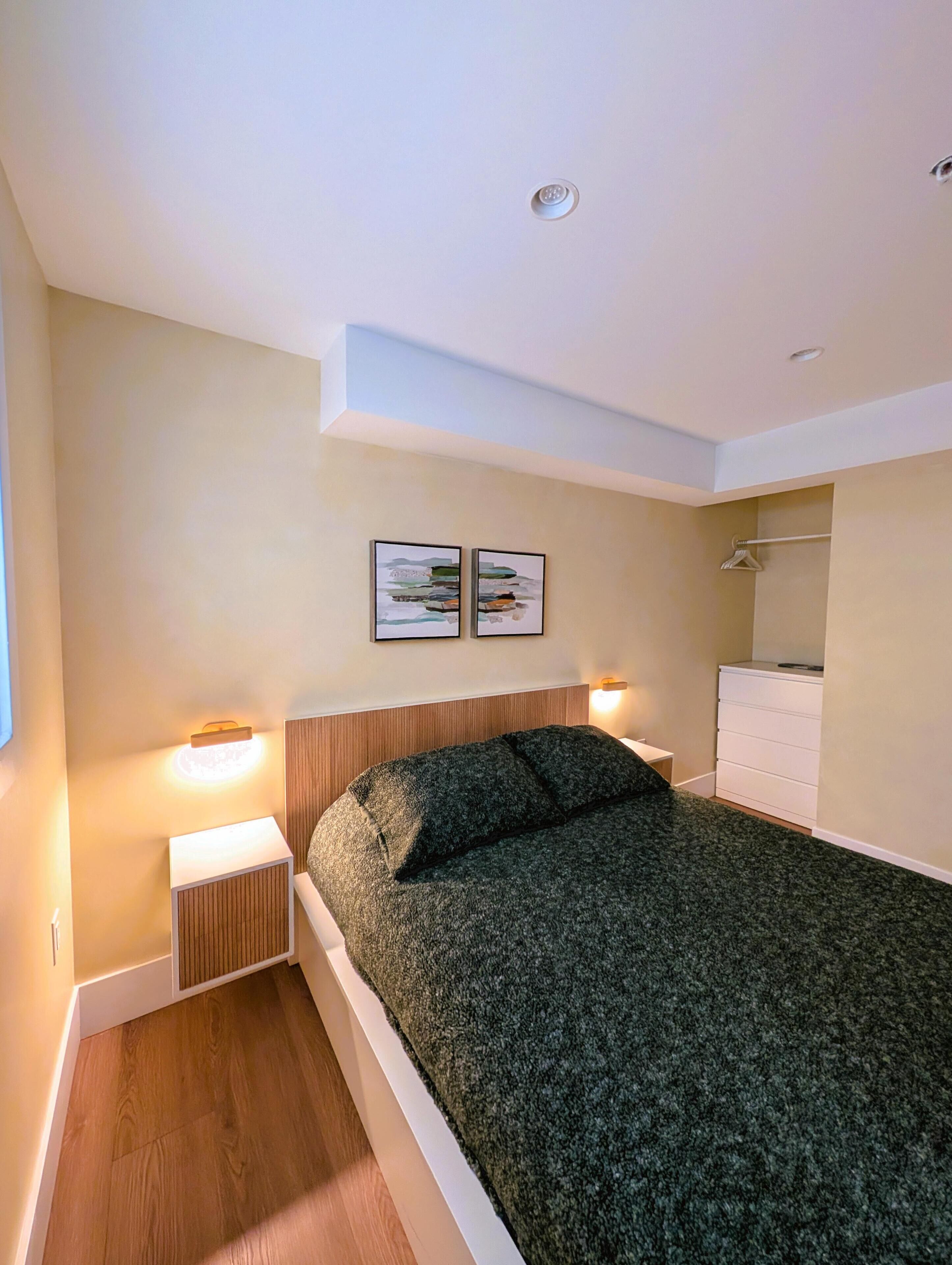 A modern bedroom with a bed, two wall-mounted lights, a wooden headboard, a white dresser, and a small nightstand.