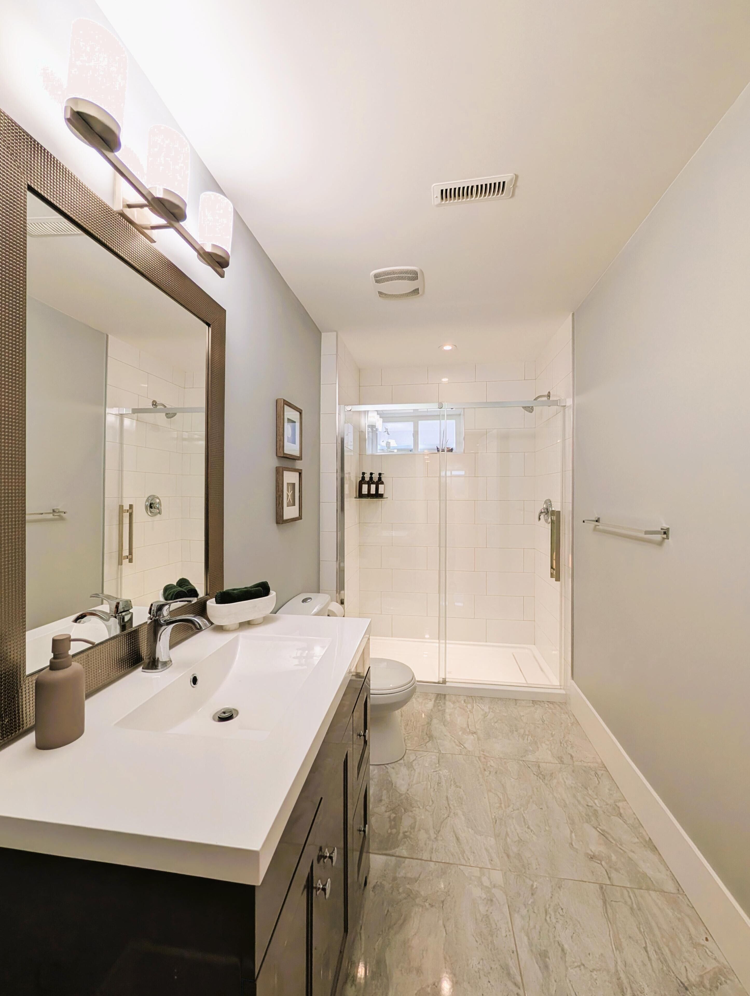 A modern bathroom with a large mirror, two sinks, a shower, and a toilet.