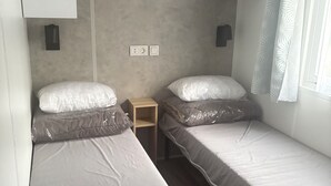 Una stanza piccola e accogliente con due letti singoli, un comodino in legno, una mensola a parete e una finestra con tenda decorata.