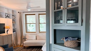 A living room with a wooden dining table, a sofa, a television, and a cabinet with shelves. - NEW 4BR Coastal Home with AC Steps to Kinney Shores Beach ⭐⭐⭐⭐⭐ (Saco)
