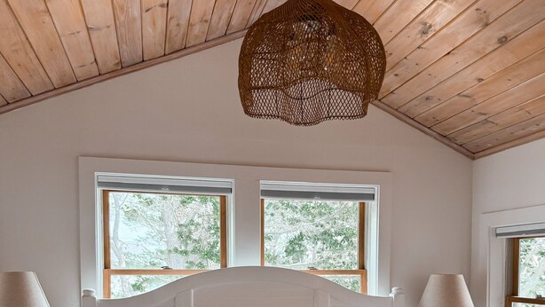 A bedroom with a wooden ceiling, a large bed with white bedding, two bedside tables with lamps, and windows with a view of trees. - NEW 4BR Coastal Home with AC Steps to Kinney Shores Beach ⭐⭐⭐⭐⭐ (Saco)