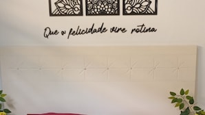 Una cama con cabecera blanca, almohadas rojas y colcha negra. Hay cuadros en la pared sobre la cama y un florero con flores en la mesita de noche.