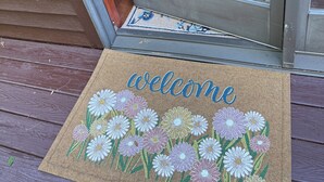 A wooden deck with a glass door entrance, a welcome mat with floral designs, and a wooden bench in the background. - Spacious 1-bedroom suite surrounded by nature in charming Downingtown. (Downingtown)