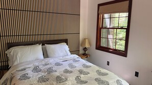A bedroom with a bed, a window with a view, a bedside lamp, and a wall with vertical lines. - Spacious 1-bedroom suite surrounded by nature in charming Downingtown. (Downingtown)