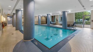 A modern indoor swimming pool with tiled walls and ceiling, clear pool area, and a view of the outdoors through large windows. - Downtown Loft-style Condo | Pool, Gym & Balcony (Toronto)