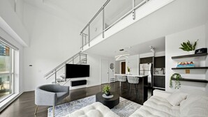 A modern living room with a sofa, a chair, a coffee table, a TV, a staircase, and a kitchen area. - Downtown Loft-style Condo | Pool, Gym & Balcony (Toronto)