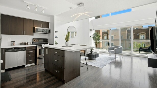 A modern kitchen with dark cabinetry, a central island, and a dining area with a view of the city. - Downtown Loft-style Condo | Pool, Gym & Balcony (Toronto)