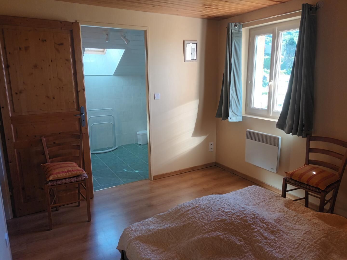 Un dormitorio con cama, dos sillas, una ventana con cortinas, un radiador y una puerta que da al baño.