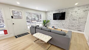 A modern living room with a grey sectional sofa, a white ottoman, a flat-screen TV mounted on a brick wall, and a large window with a view of trees. - Modern Lakefront Escape In Muskoka (Emsdale)