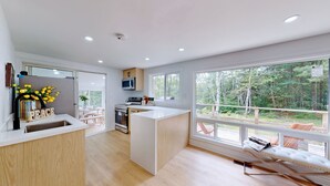 A modern kitchen with a central island, a dining area with a bench, and large windows offering a view of a forest. - Modern Lakefront Escape In Muskoka (Emsdale)