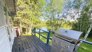 A barbecue grill on a wooden deck with a railing, surrounded by trees and a clear sky. - Modern Lakefront Escape In Muskoka (Emsdale)
