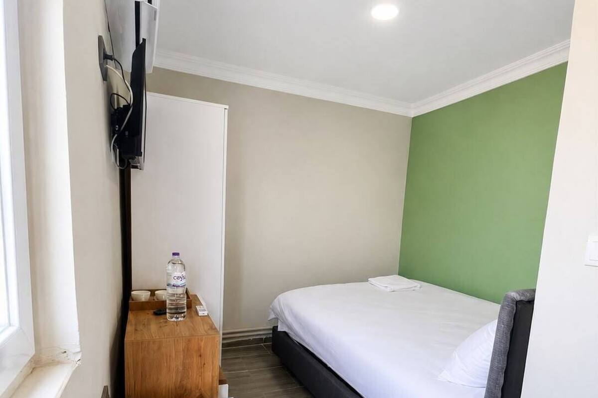 Una habitación de hotel con una cama, una pequeña mesa de madera con una botella de agua, un cubo de basura blanco y un equipo de aire acondicionado instalado en la pared.
