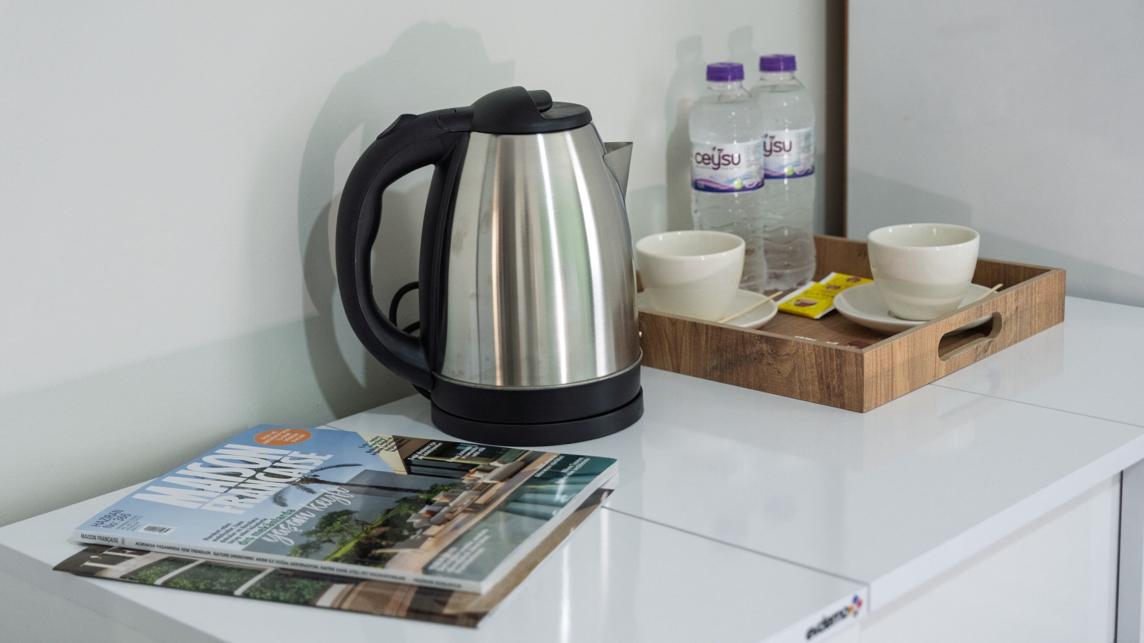 A stainless steel electric kettle, two water bottles, a tray with two white cups and saucers, and a magazine on a white surface.