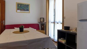 Une pièce comprenant un lit, une table avec un set de table, une plante, une bibliothèque remplie de livres, un tableau encadré au mur et une fenêtre avec des rideaux.