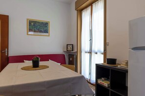 A room with a bed, a table with a placemat, a plant, a bookshelf with books, a framed picture on the wall, and a window with curtains. - Appartamento Magnolia - Affitti Brevi Italia (Borghetto Santo Spirito)
