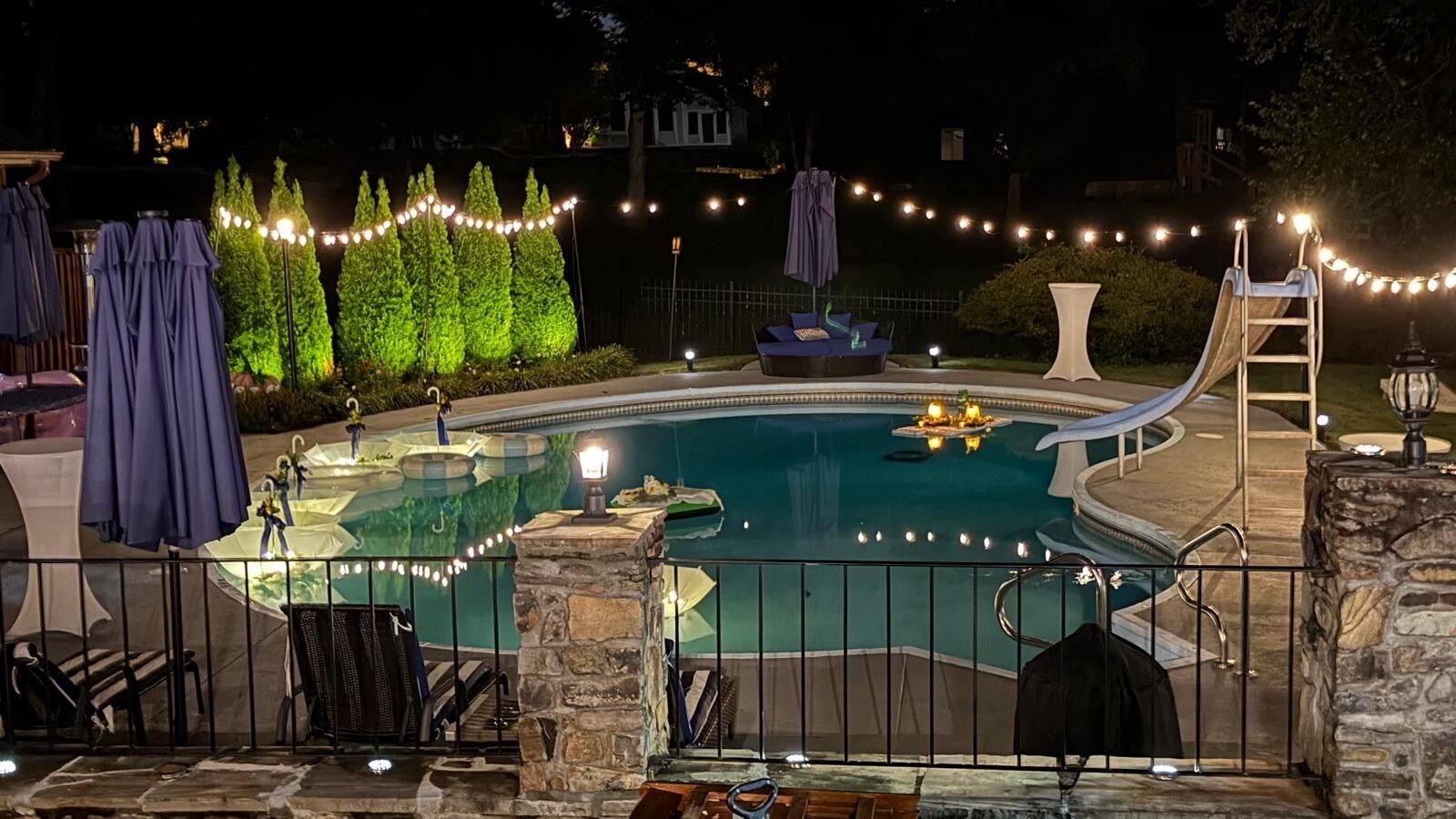 Une piscine bordée d’un muret en pierre, avec des chaises longues et des guirlandes lumineuses.