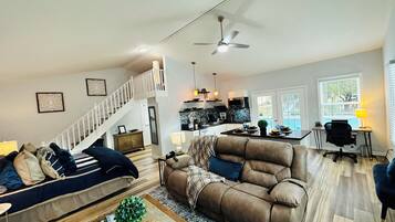 Un vaste espace de vie comprenant un canapé, une table basse agrémentée d’objets décoratifs, une table à manger avec des chaises, un coin cuisine et un escalier.